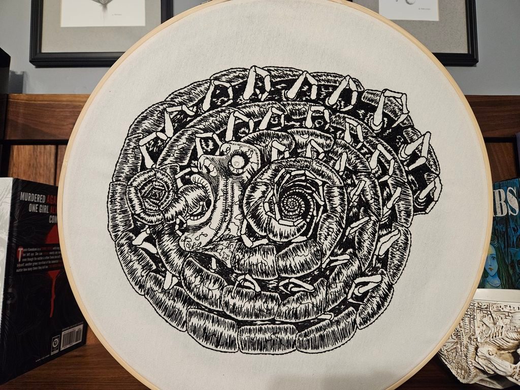 Photo of an embroidery hoop containing the black and white embroidered image of the centipede man by Junji Ito. It's a coiled centipede with the face of a man growing out of its body, a horrified look on his face as his tongue coils out of his gaping mouth.